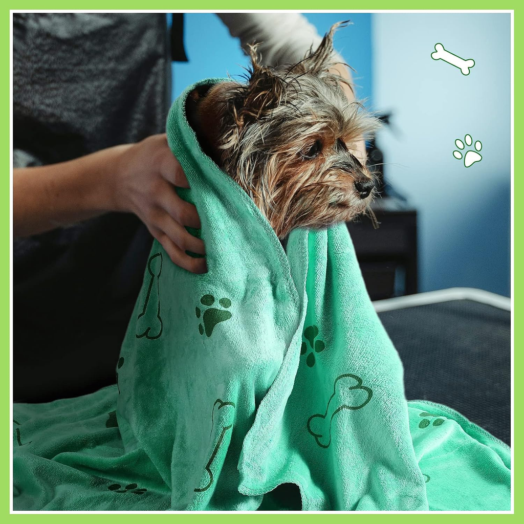 A small wet dog wrapped in a mint green towel with dark green bone and paw print patterns, held by a person in a grooming setting.