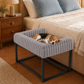 A small dog rests on a gray ribbed pet bench with a black metal frame, placed beside a bed in a warmly lit bedroom.